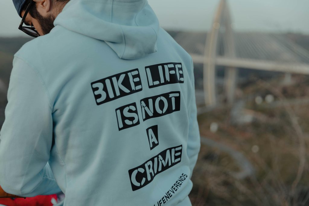 Man in gray hoodie with message 'Bike Life Is Not A Crime' outdoors near a bridge.
